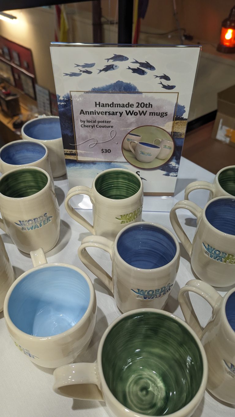 Ceramic mugs with blue and green interiors surround a sign that reads "Handmade 20th Anniversary WoW mugs by local potter Cheryl Couture, $30," displayed on a white tablecloth.