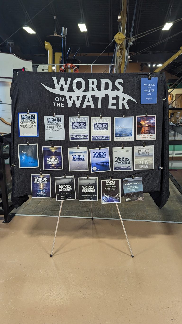 A black display board with "Words on the Water" at the top, showcasing multiple event posters with text and images, set up indoors on a tripod stand.
