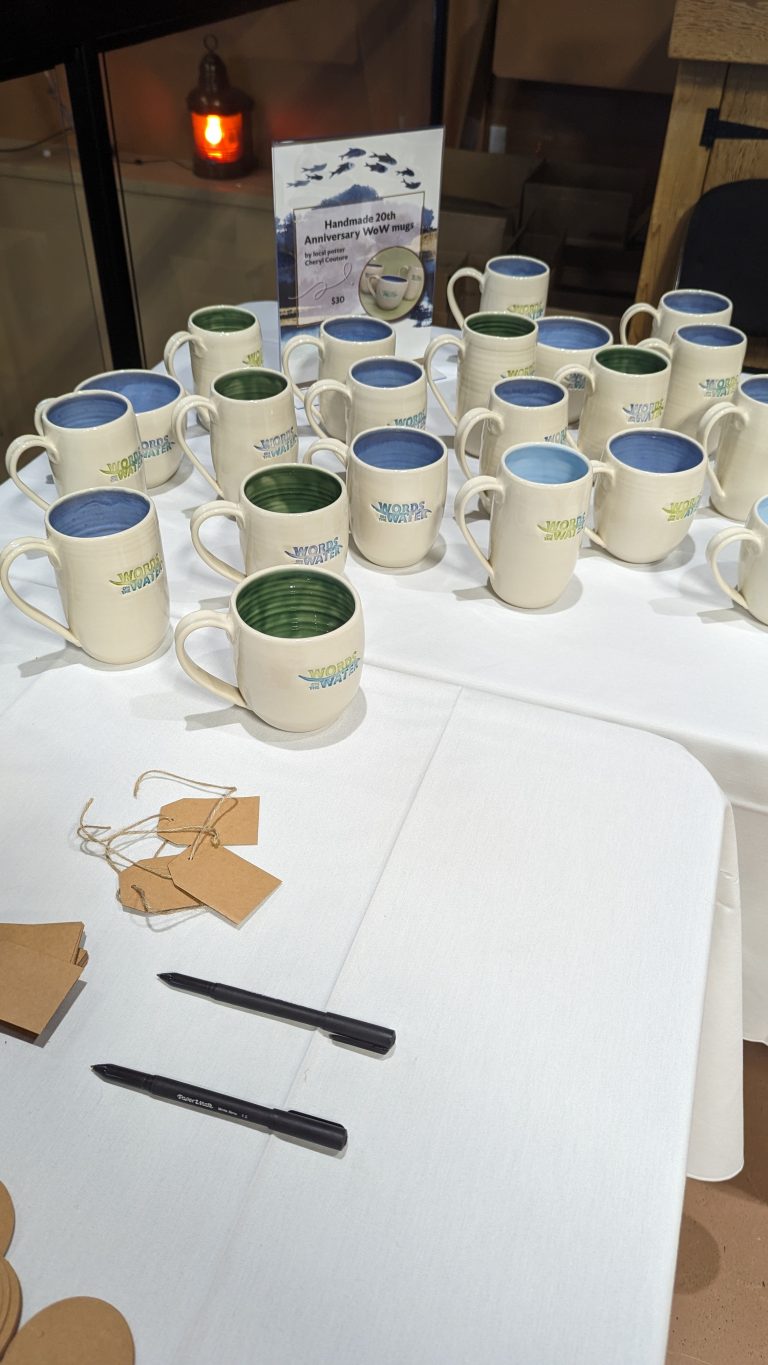 Twenty ceramic mugs with colored interiors are displayed on a white tablecloth. Two pens, blank tags, and a small sign with event details are also on the table. A lit candle is visible in the background.