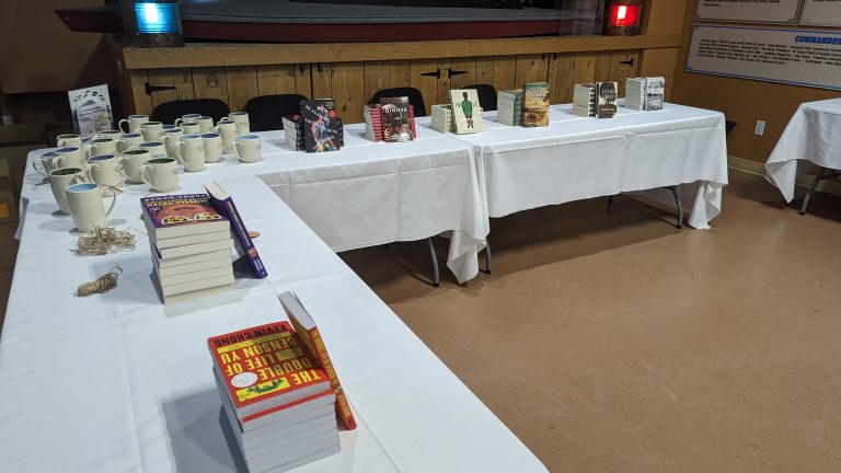 Tables with white tablecloths display mugs, several stacks of books, and some jewelry. There are chairs in the background and a wooden partition behind the tables.