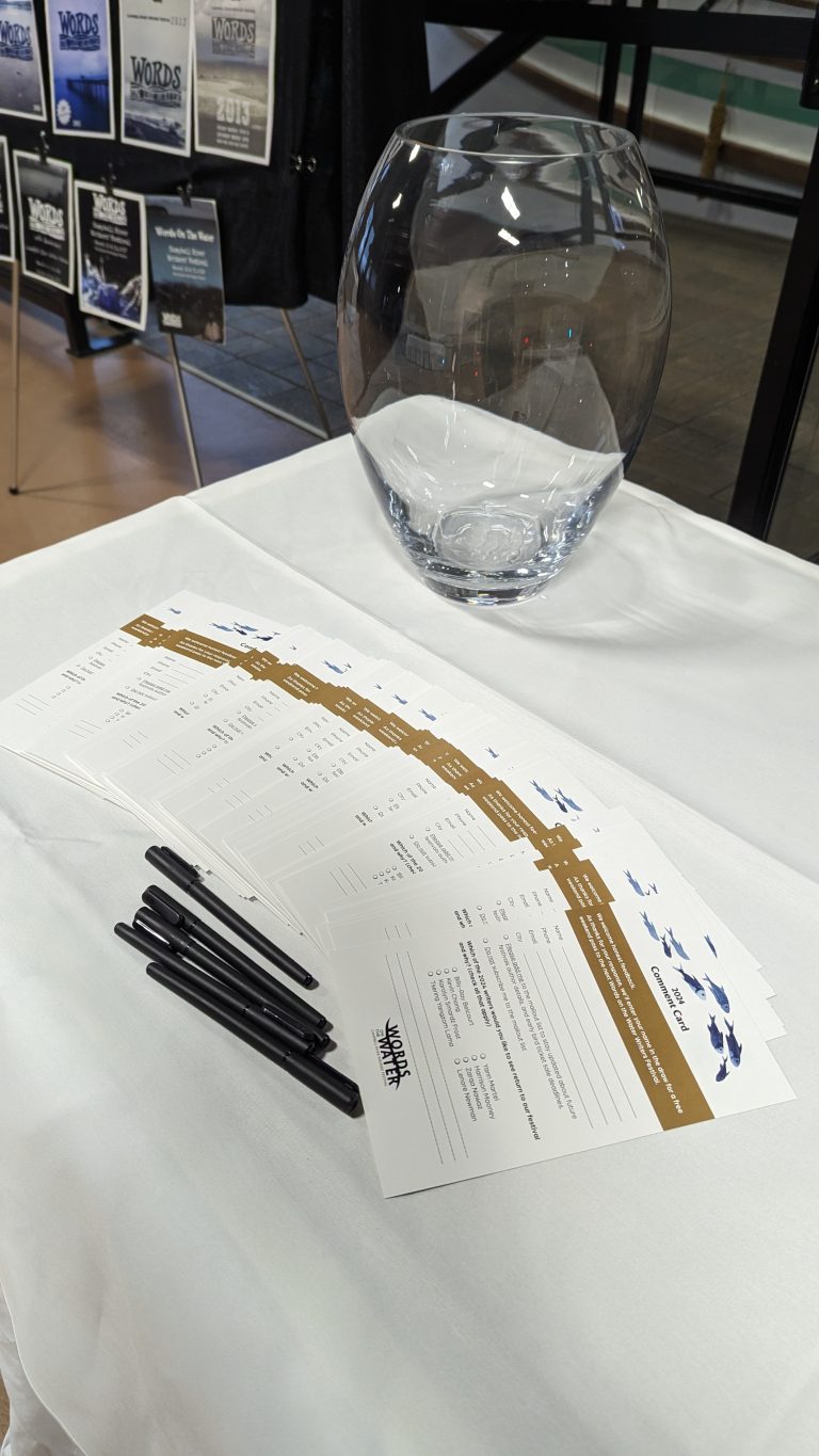 A bowl and several black pens are on a white tablecloth with forms titled "Words on the Wing" for an event. Posters are visible on a display board in the background.