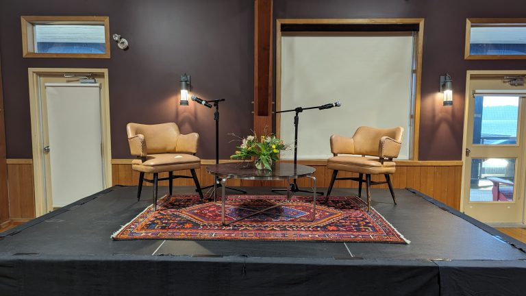 Two tan chairs face each other on a small stage with microphones, a round table with a flower arrangement, and a colorful rug. The backdrop includes brown walls, windows, and two wall lights.