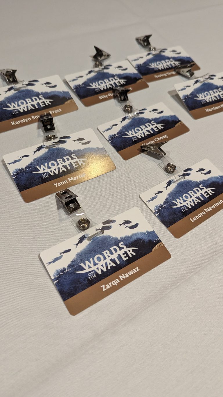 Eight name badges with "Words on Water" logo and mountain design are arranged on a white table, each with a metal clip and different names, prepared for an event or conference.