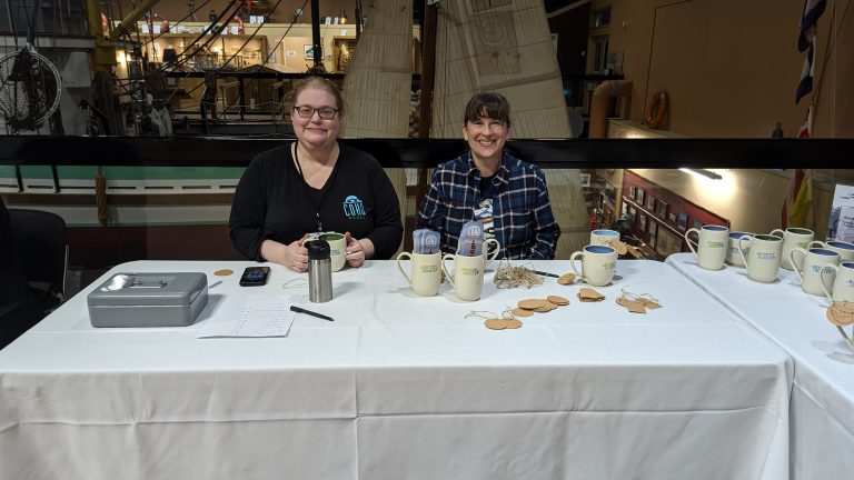 Two women sit behind a white-clothed table at a maritime museum, with mugs and craft supplies displayed. The woman on the left wears a black "Coho Books" shirt, and the woman on the right wears a blue plaid flannel shirt.
