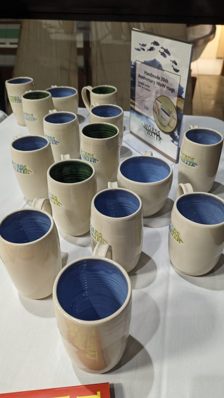 A display of ceramic mugs, mostly white with blue or green interiors, arranged on a round white tablecloth. A sign in the back reads “Handmade 20th Anniversary WOW mugs.”.