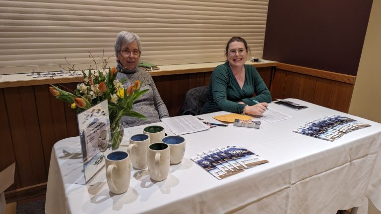 Two women sit at a table with brochures, mugs, flowers, and sign-up sheets. Both are smiling, and the table is set up in a warmly lit indoor space with wood-paneled walls.