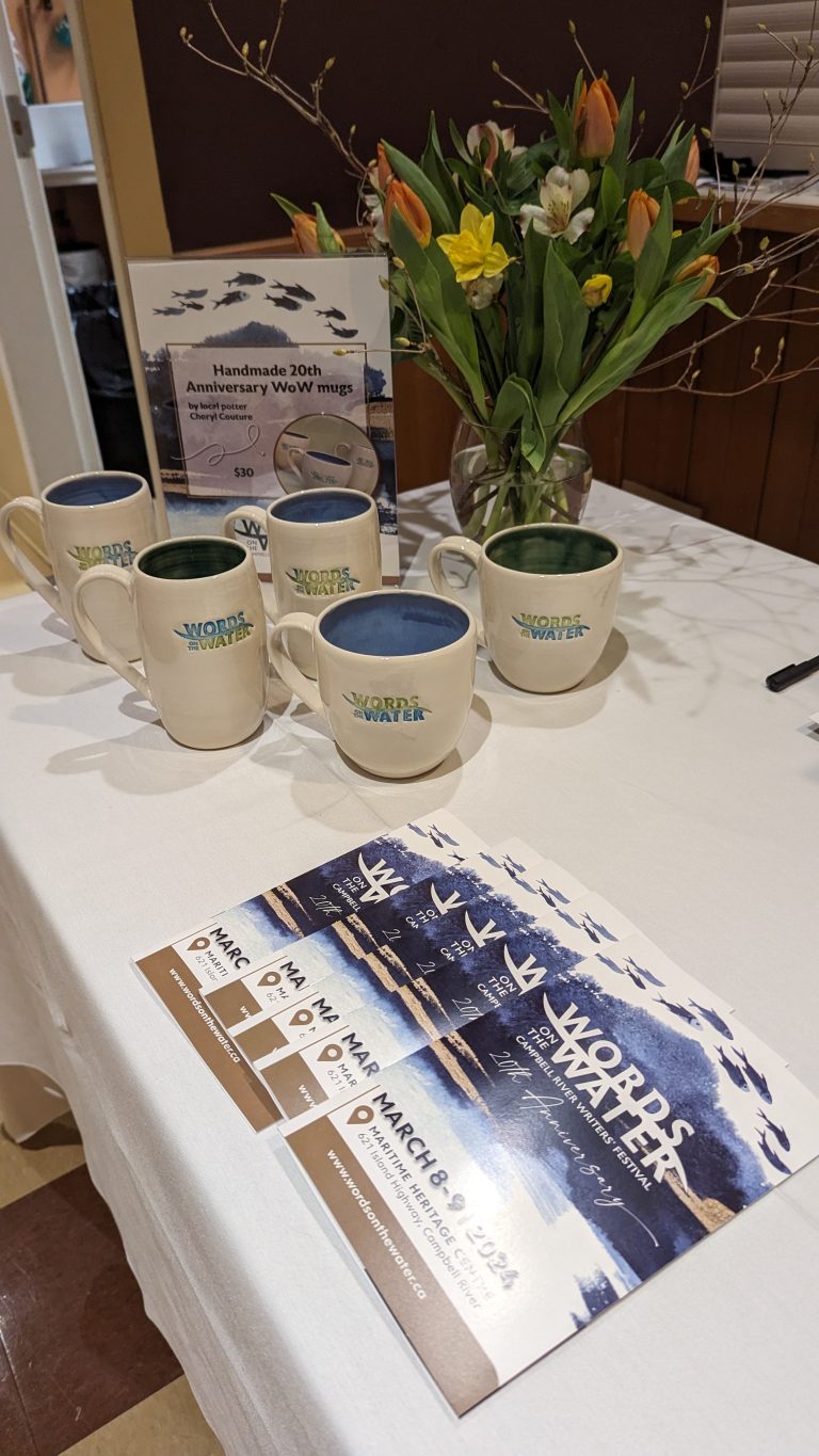 Table with white cloth displaying four handmade "Words on the Water" ceramic mugs. A sign advertises them for $30, next to a vase of tulips and daffodils. Several event brochures for the 20th Anniversary festival are laid out on the table.