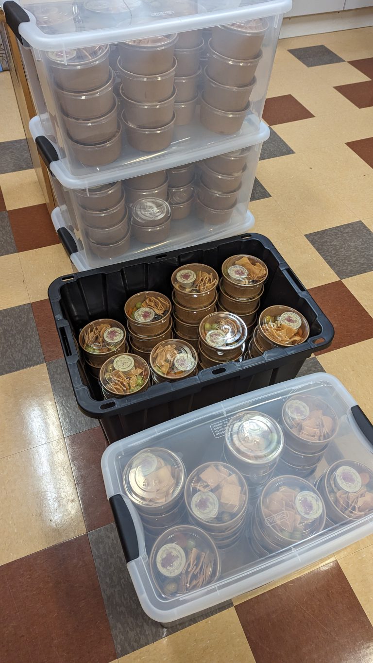Several clear and black storage bins filled with stacked, sealed food containers sit on a checkered tile floor. The food containers appear to hold individually packed meals or snacks.