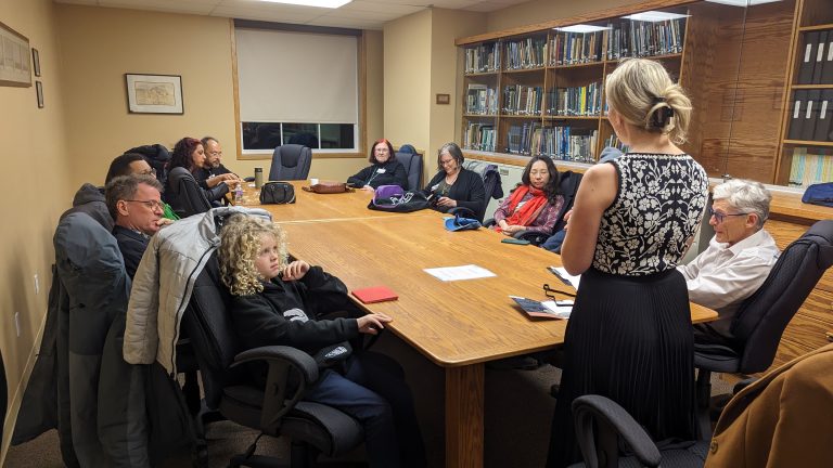 A group of people sit around a large table in a library meeting room, listening to a woman standing and speaking. Bookshelves line the wall, and papers are on the table.