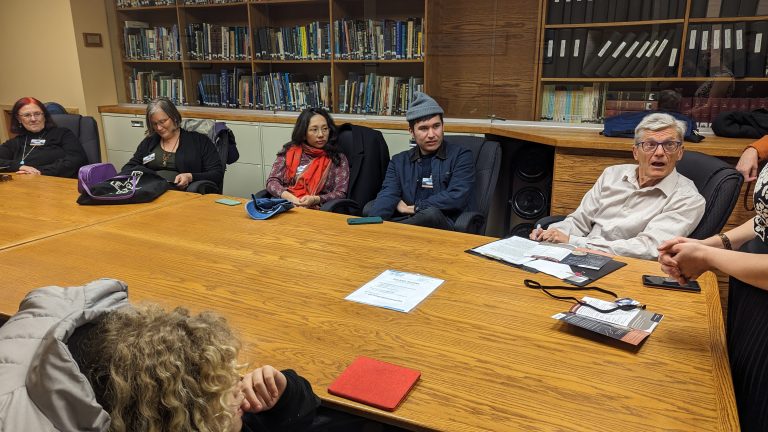 Seven people sit around a wooden conference table in a library or office, engaged in conversation. Papers, a bag, and other items are on the table. Bookshelves are in the background.