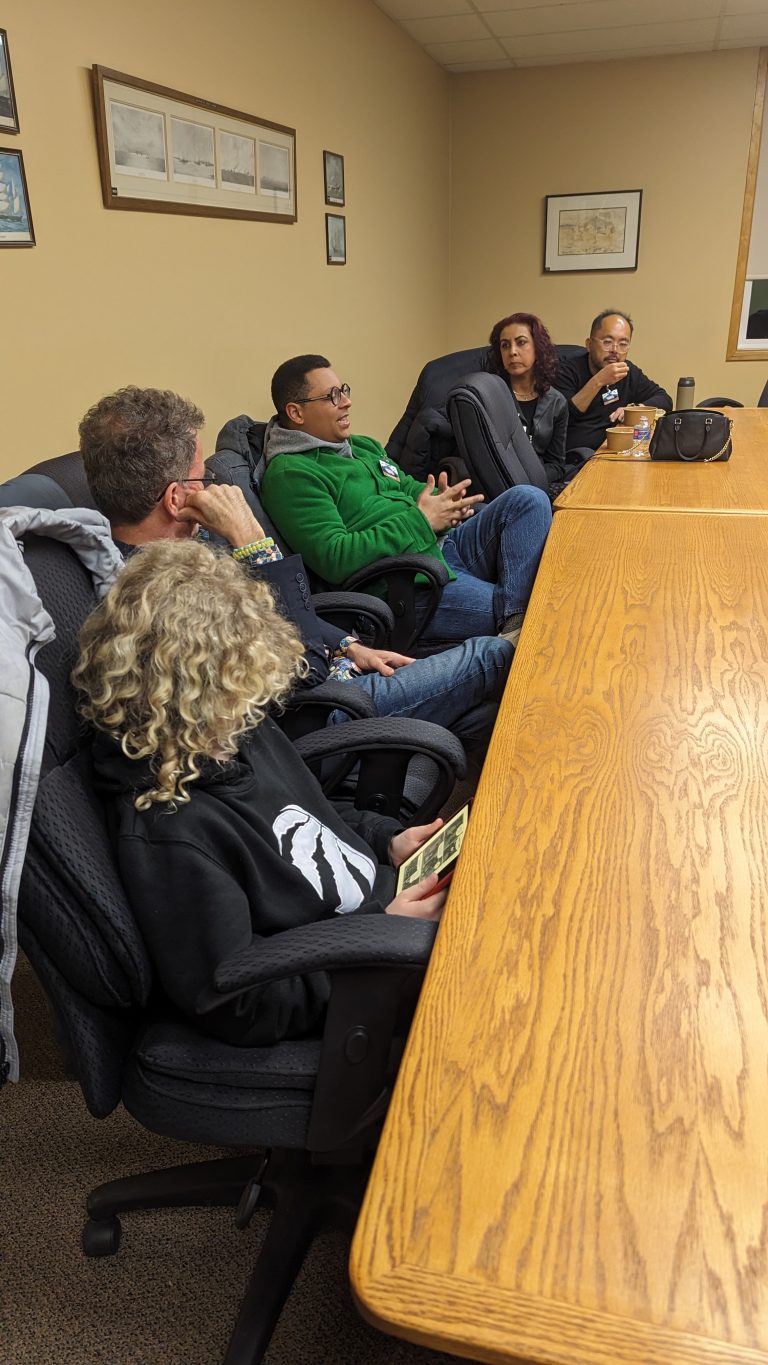 Five people sit around a conference table engaged in discussion. One person in a green hoodie gestures while speaking, while others listen. A child with curly blonde hair holds a card or notepad.