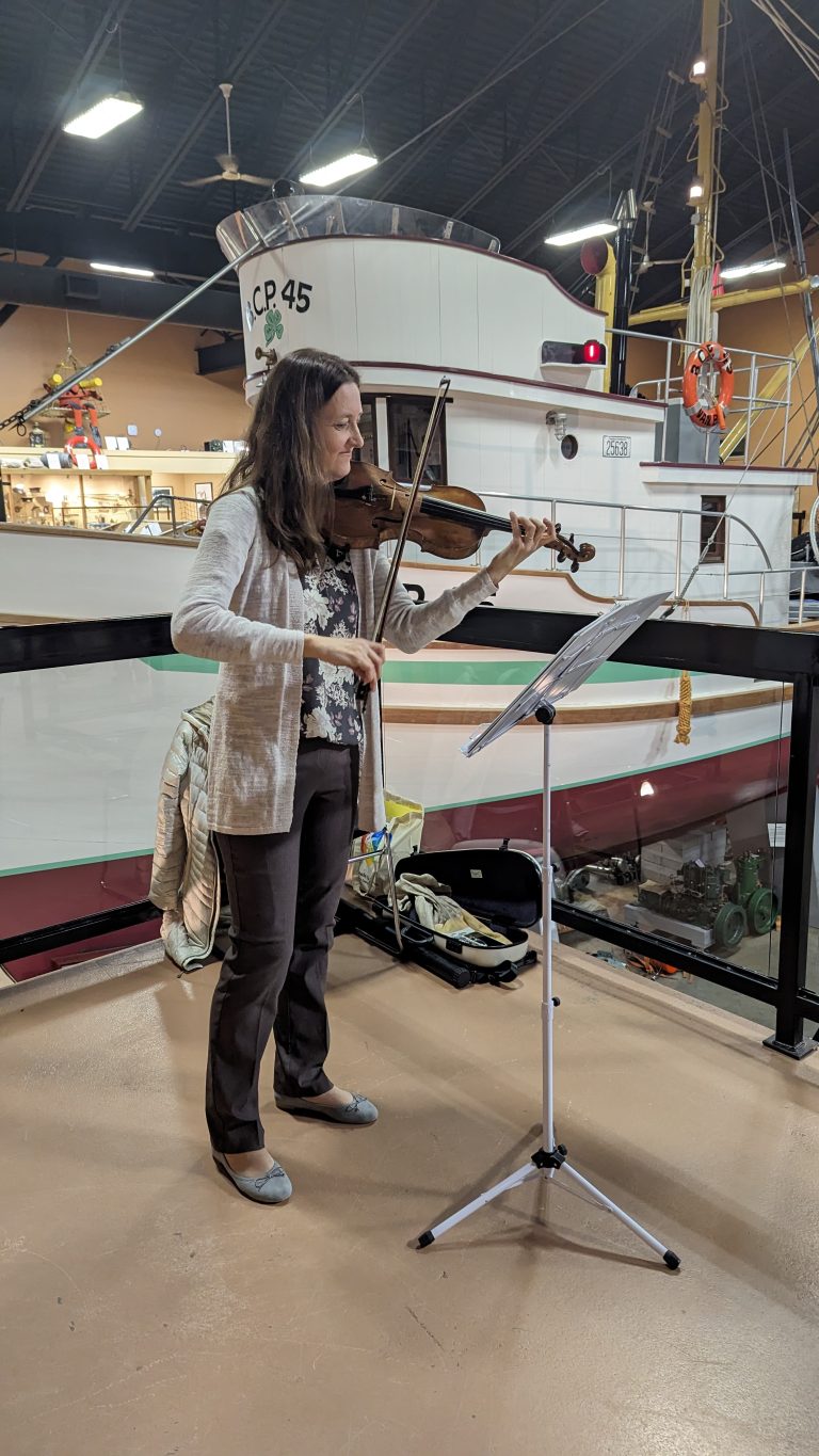 A woman plays the violin, reading sheet music on a stand, inside a maritime museum with a large fishing boat exhibit in the background.