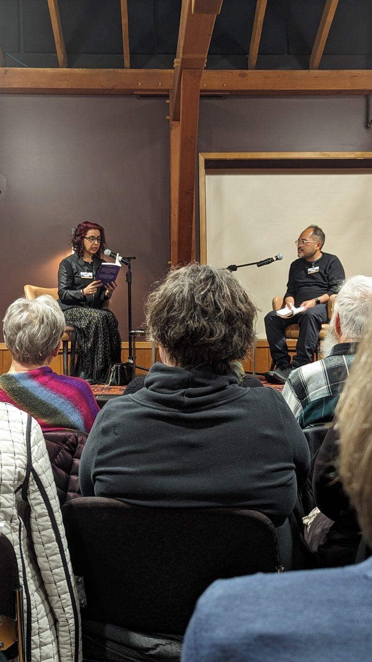 Two people sit on stage with microphones, one reading from a book, while an audience listens. The room has wooden beams and a blank screen in the background.