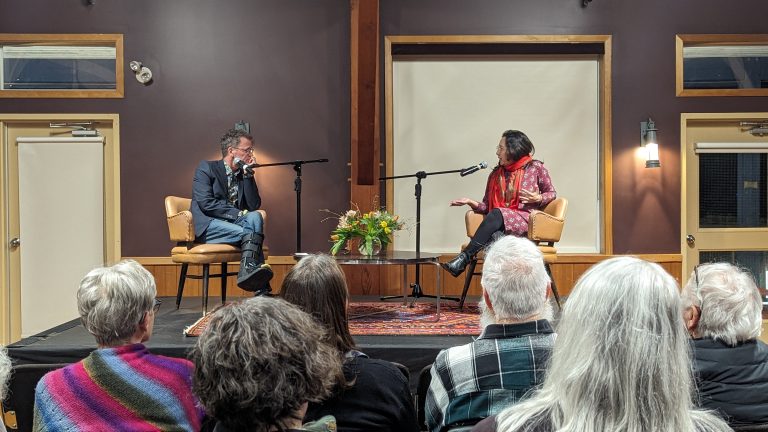 A man in a suit and a woman in a patterned top sit on stage in chairs facing each other, speaking into microphones. A small audience watches from the foreground in a room with dark purple walls, wood trim, and a patterned rug on the stage.