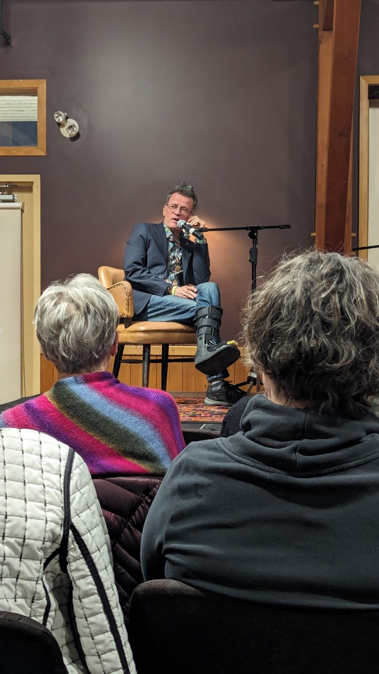 A person in a suit sits on a chair, holding a microphone and speaking. Two people with gray hair sit in the audience, one wearing a colorful shawl. The setting appears to be a small indoor event or talk.