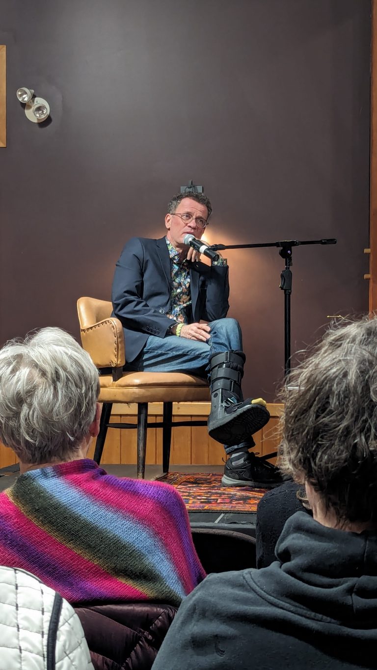 A man in a blazer, jeans, and a leg brace sits on a chair, speaking into a microphone. He is on a small stage facing an audience. The room has dark walls and warm lighting.