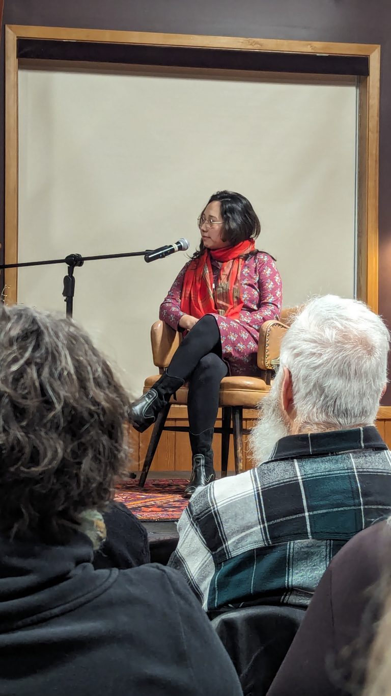 A woman in a red scarf and patterned top sits cross-legged on a chair, speaking into a microphone on stage, while audience members listen attentively.
