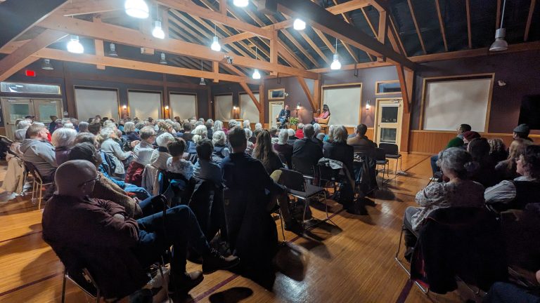 A large audience sits in a wooden-beamed hall, attentively watching a woman speak at a podium under warm lighting.