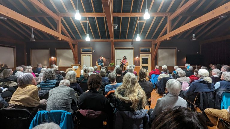 A large group of people sits in rows facing a stage where two individuals stand and speak inside a wooden-beamed hall with bright ceiling lights.