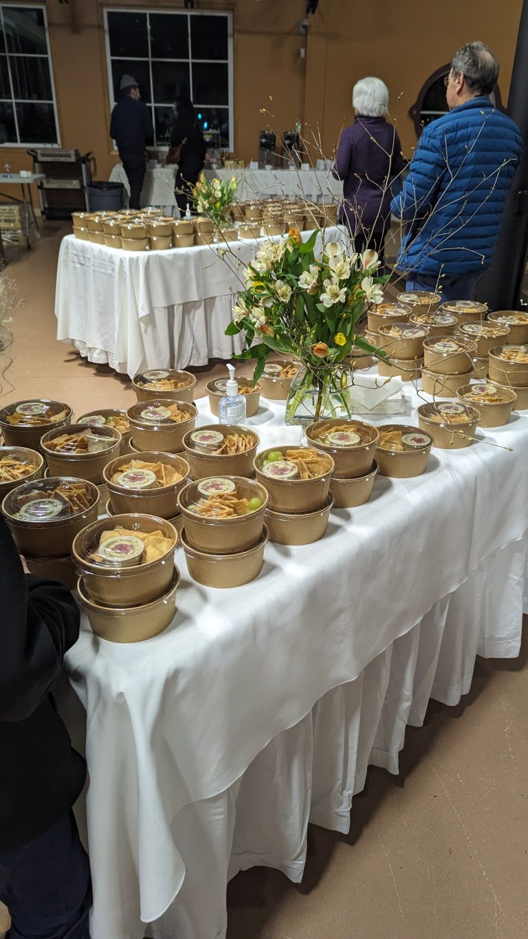 Stacked food containers on tables covered with white cloths, with flowers as centerpieces. Several people are standing and walking around in the background. Indoors, possibly at a catered event or banquet.