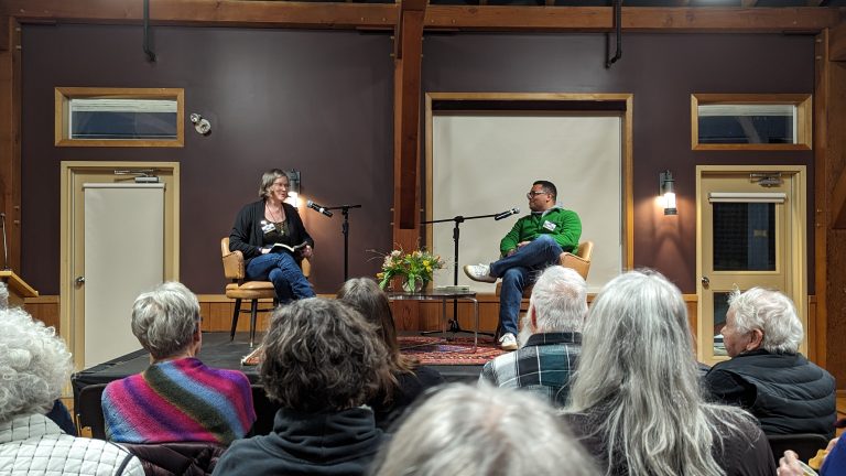 A woman and a man sit in armchairs on a small stage, speaking into microphones for an audience. There is a small table with a floral arrangement between them, and the room has wood-paneled walls and dark brown accents.