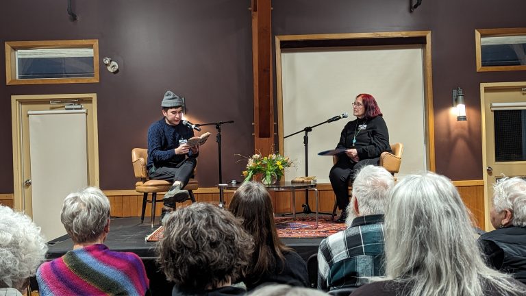 Two people sit on stage with microphones, one reading from a book. There’s a flower arrangement on a table between them. An audience watches, and the room has dark walls with beige doors and windows.