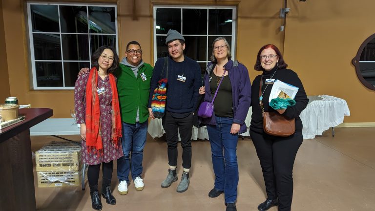 Five people stand indoors smiling at the camera; three women and two men in casual clothes. A table with a white cloth and windows are in the background. One person holds a small, colorful knitted item.