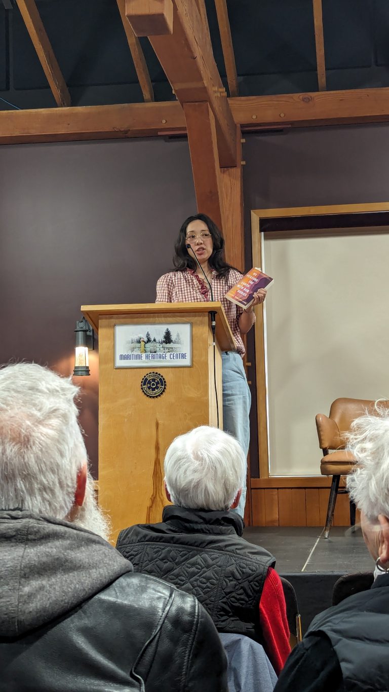A woman stands at a podium speaking into a microphone, holding a book. She faces an audience seated indoors. The podium sign reads “Maritime Heritage Centre.” The room has wooden beams and a deep purple wall.