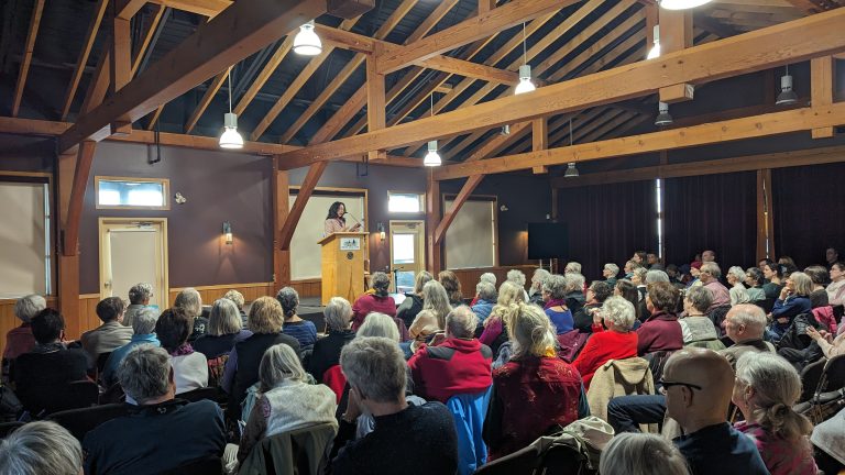 A woman speaks at a podium in a wooden hall, addressing a large seated audience. The attendees are mostly older adults, attentively listening under exposed beams and warm lighting.