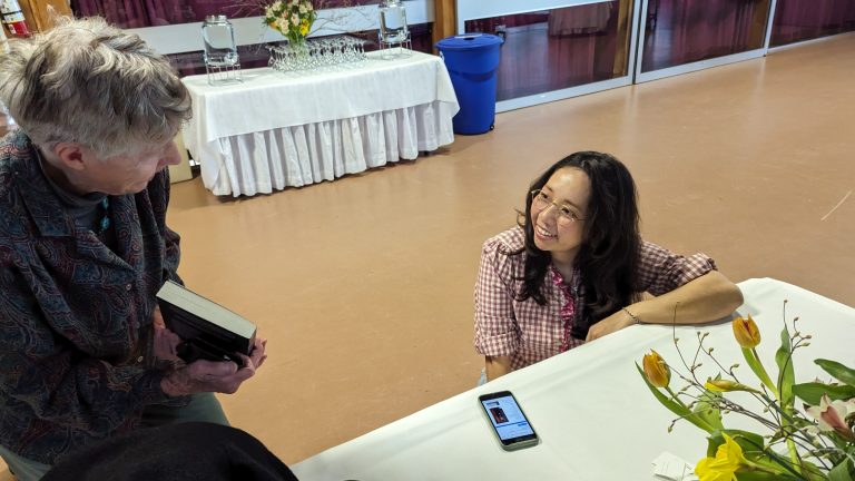 A smiling woman sits at a table with flowers and a phone, talking to an older person holding books. A table with glasses and a blue bin are in the background.