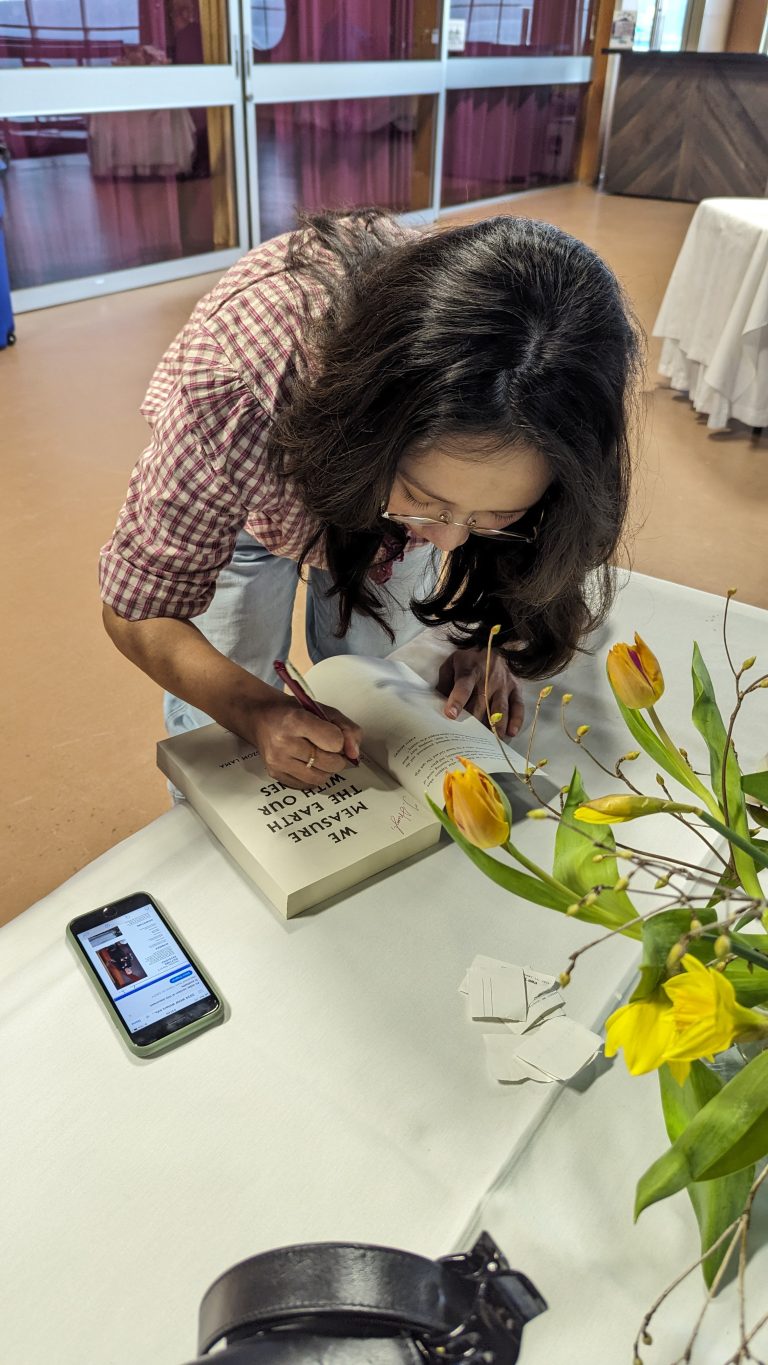 A person with long dark hair signs a book at a table with yellow flowers. A smartphone displaying a webpage and some paper scraps are also on the white tablecloth.