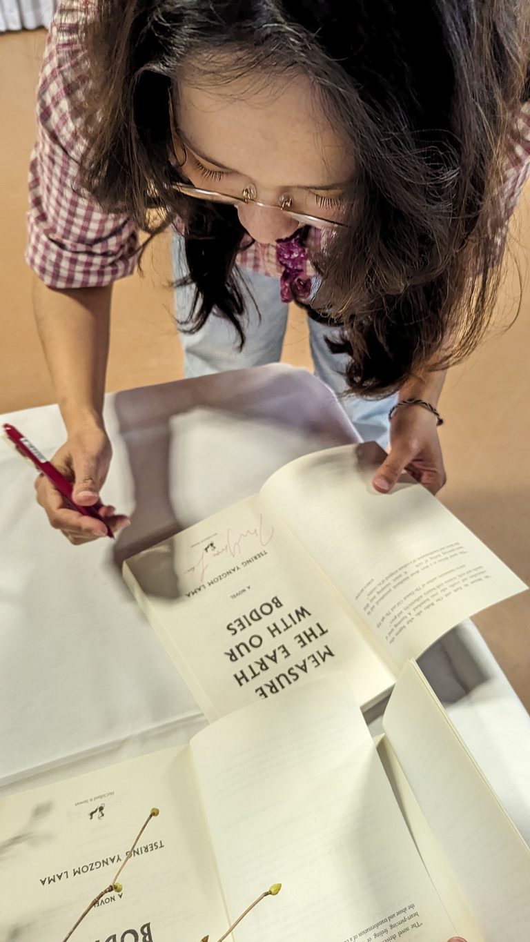 A woman with long hair and glasses signs a book with a red pen while standing at a table, looking down at the open pages. She wears a plaid shirt and appears focused on her task.