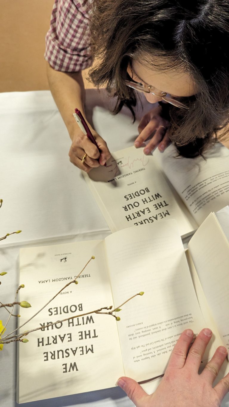 A woman with glasses signs a copy of the novel "We Measure the Earth with Our Bodies" while another person holds the book open on a table with yellow flowers.