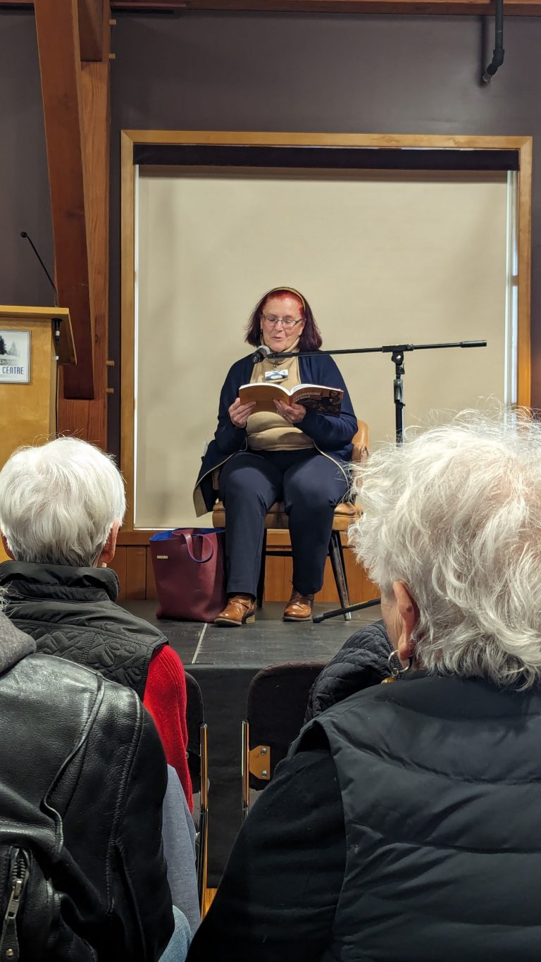 A woman sits on stage reading from a book to an audience. She wears glasses and a red headband. Several people with gray hair are seated in front of her, listening. A microphone is set up nearby.