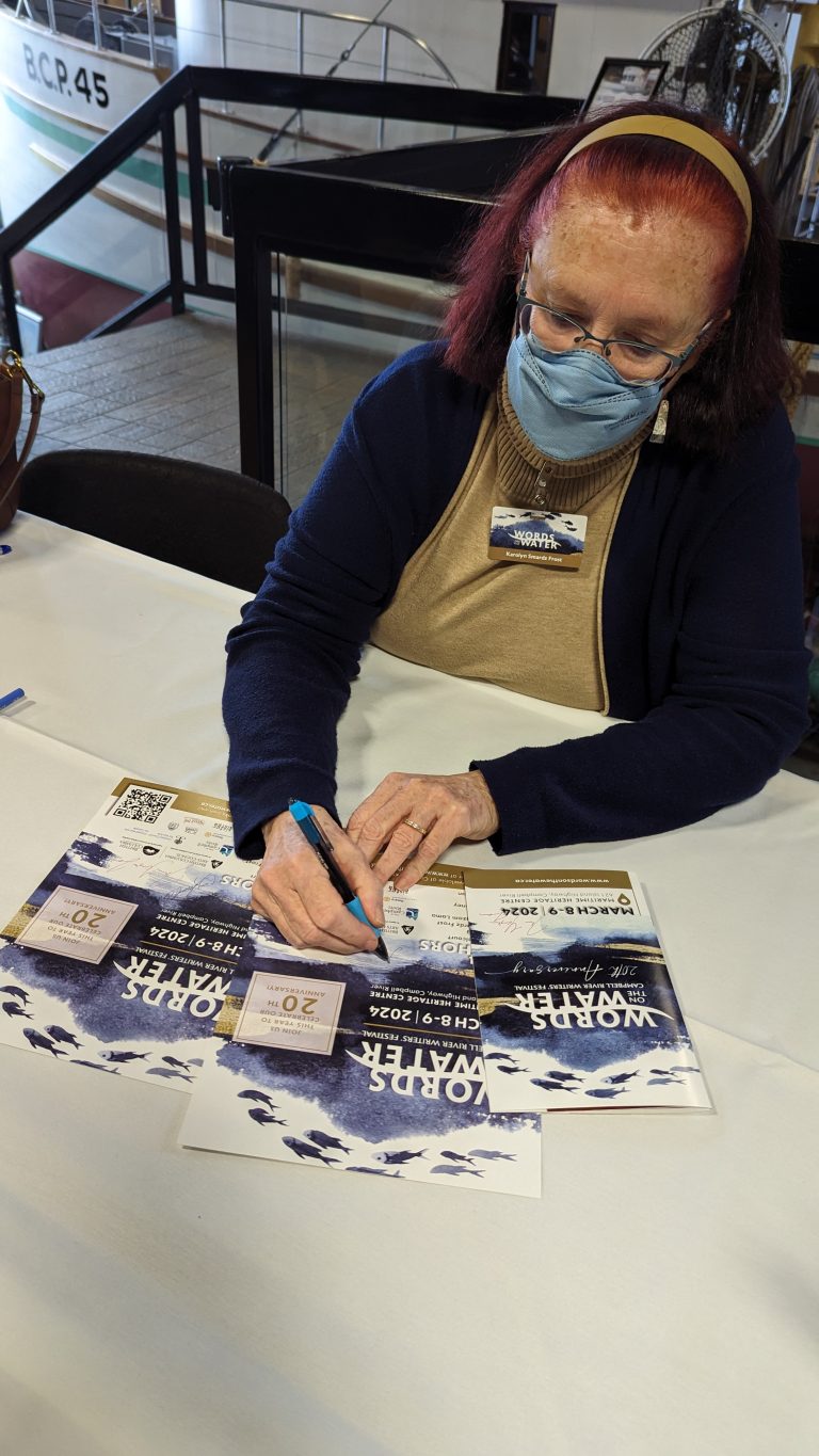A woman with red hair, glasses, and a blue mask sits at a table signing multiple flyers for an event called "WORK CYFAIR" with a blue marker.