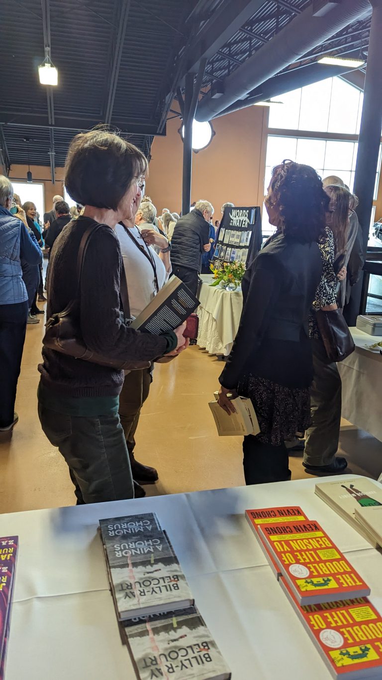 People gather at a book event indoors. Two women in the foreground talk while holding books. A table with books is visible, and more attendees stand near a window in the background.