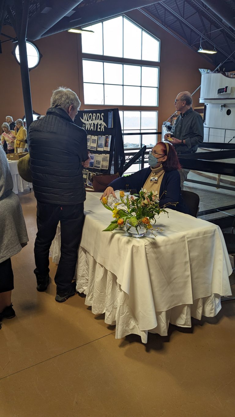 A woman sits at a table with flowers, smiling and signing books for a man. A display behind her reads "Words & Water." Other people are present in the background at the indoor event.