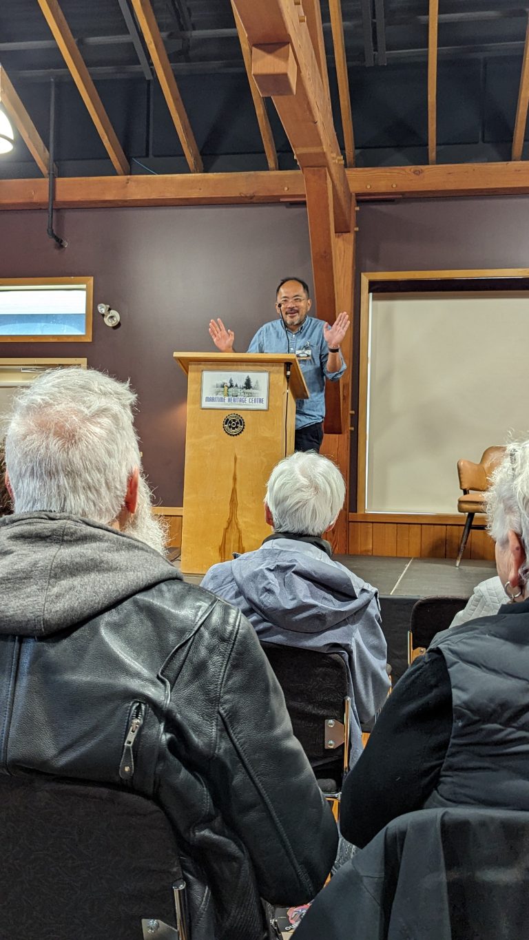 A man stands at a wooden podium, speaking to an audience in a room with exposed beams. Several seated people are visible from behind, listening attentively. The man gestures with his hands while presenting.