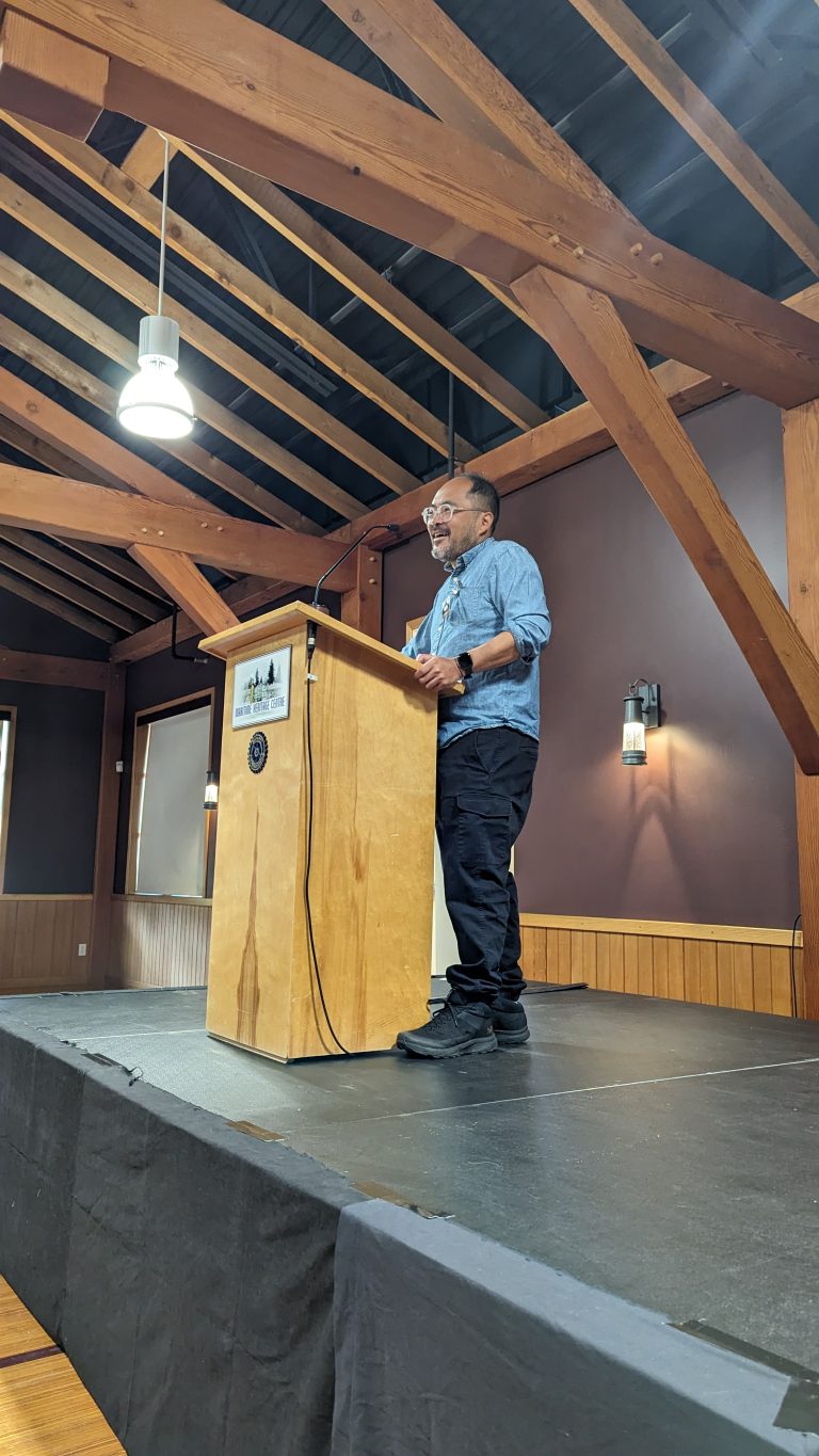 A person stands at a wooden podium on a stage, speaking in a room with exposed wooden beams and dark walls. They appear to be giving a presentation or speech.