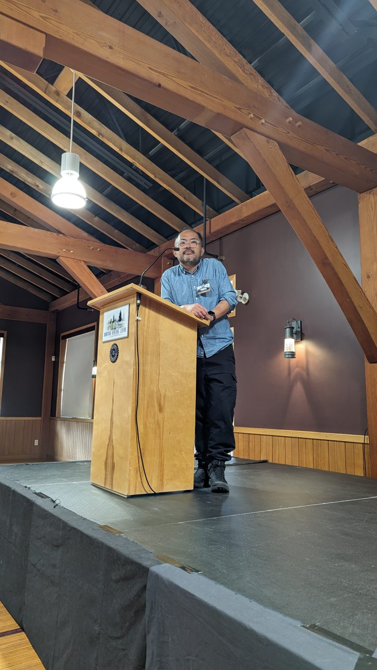 A man stands behind a wooden podium on a stage in a room with exposed wooden beams and high ceilings, speaking confidently to an unseen audience.