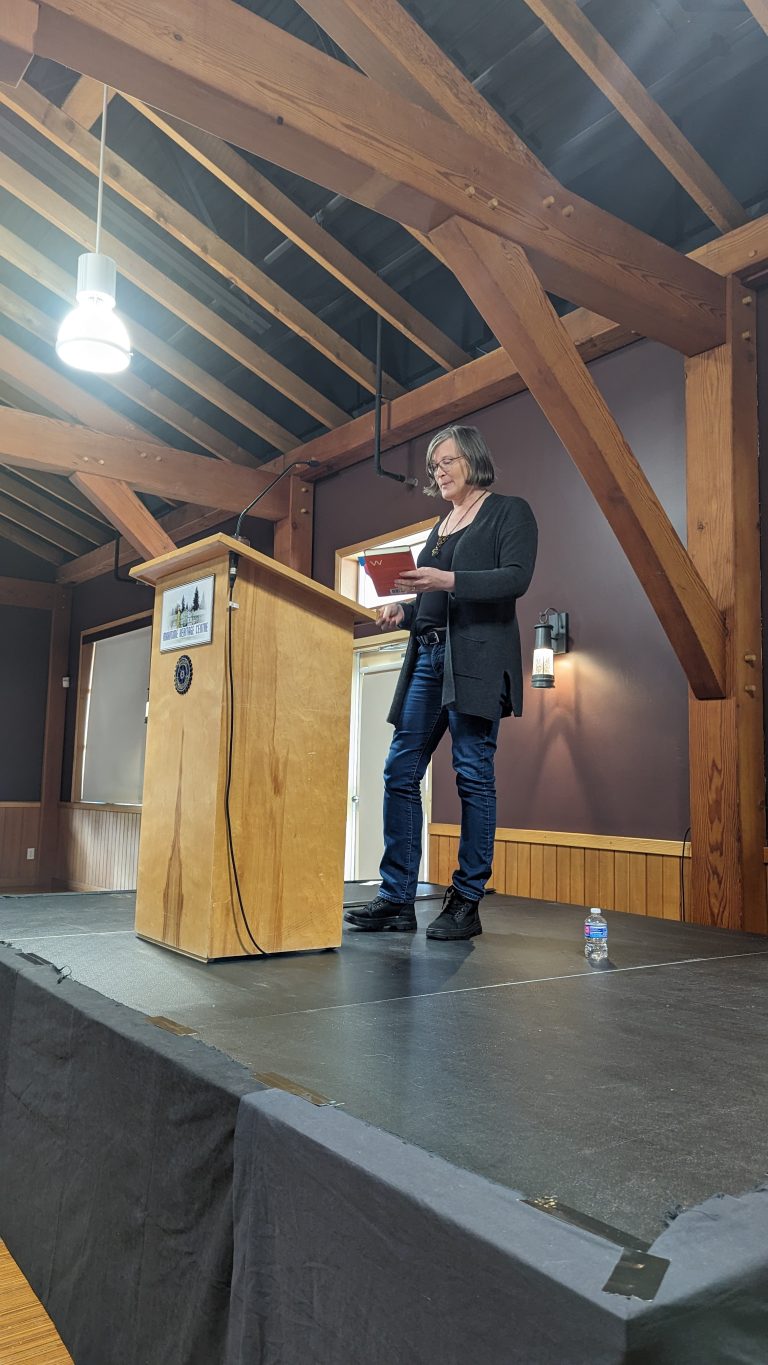 A person stands at a wooden podium, reading from a book on a stage in a room with exposed beams and purple walls. A small water bottle is on the floor beside them.