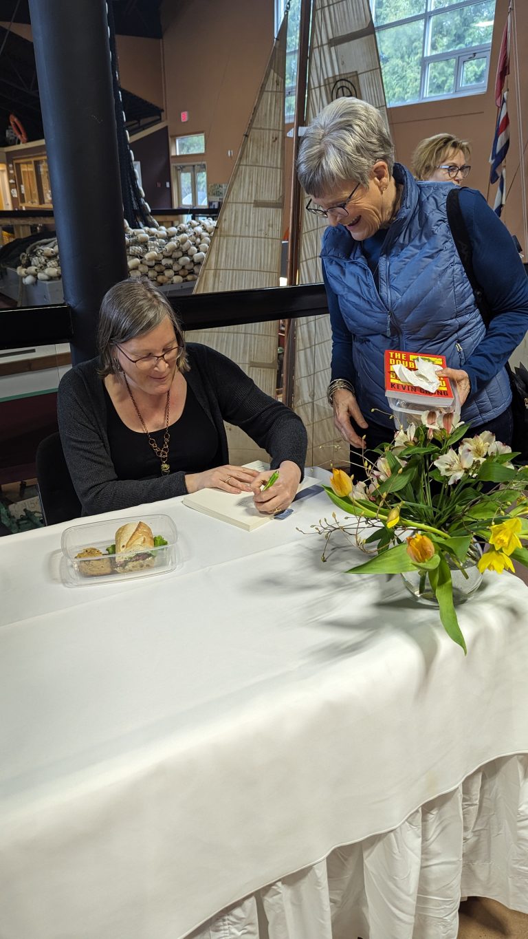 A woman sits at a table signing a book for another woman who is smiling and holding books. A plate of food and a vase with yellow flowers are on the table. A large model boat is in the background.