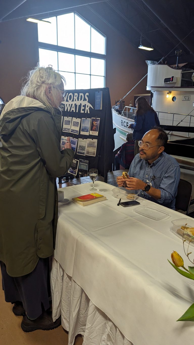 A person stands talking to an author seated at a "Words on Water" book table in a room with a boat display and large windows, with books and brochures visible on the table.