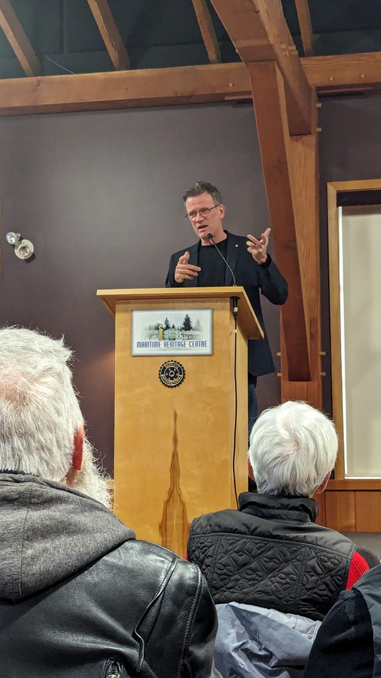 A person speaks passionately at a podium labeled "Maritime Heritage Centre" in a wooden-beamed room, while audience members listen attentively from their seats.