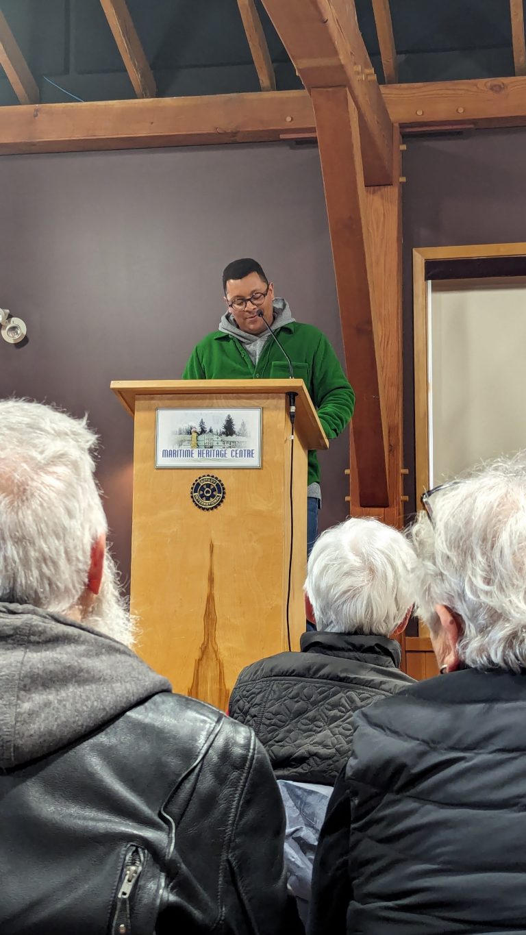 A man in a green jacket speaks at a podium in front of an audience. The podium reads "Maritime Heritage Centre." Exposed wooden beams and a brown wall are visible in the background.