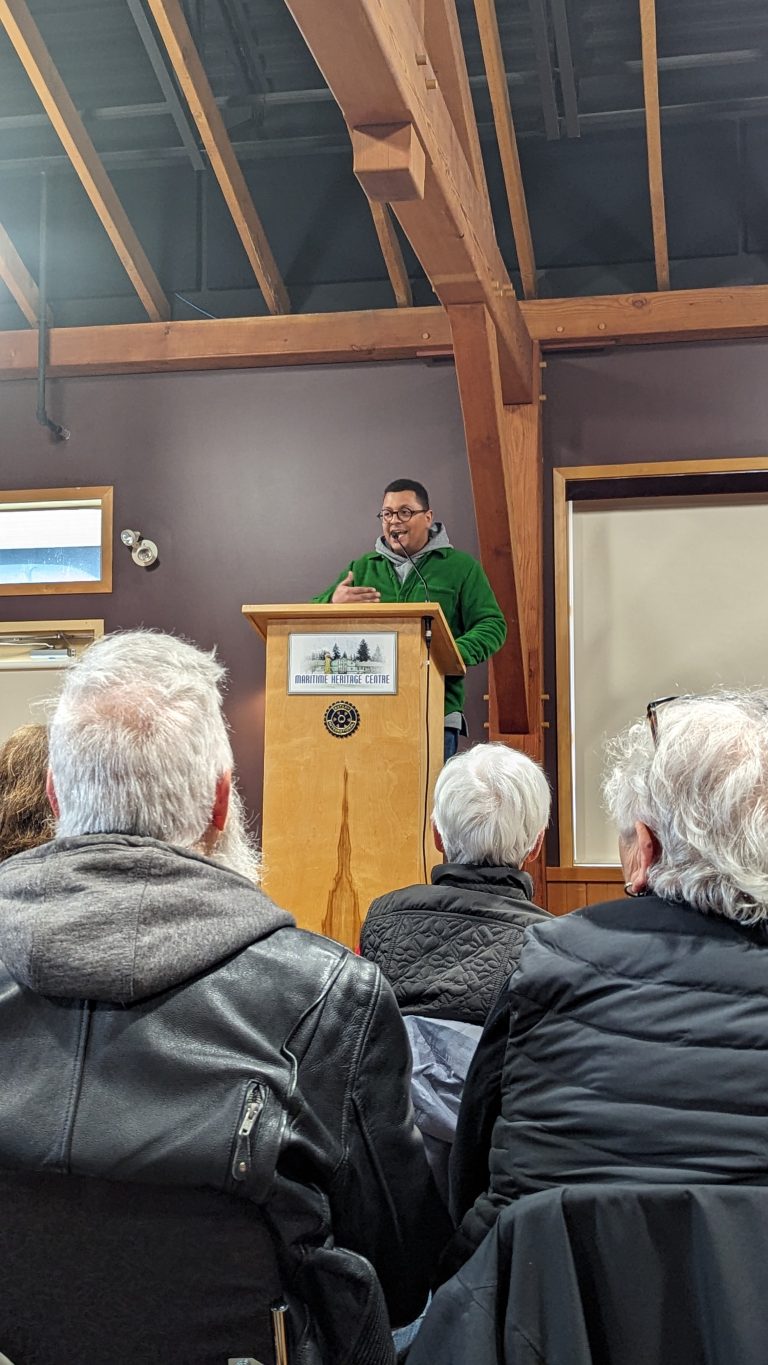 A man in a green sweater speaks at a podium in front of an audience in a wooden-beamed room. The audience is seated and listening attentively.