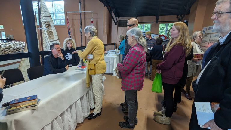 People stand in line indoors at a table, where a man appears to be signing books. Several people hold books, and others watch or wait their turn. Various objects are visible in the background.
