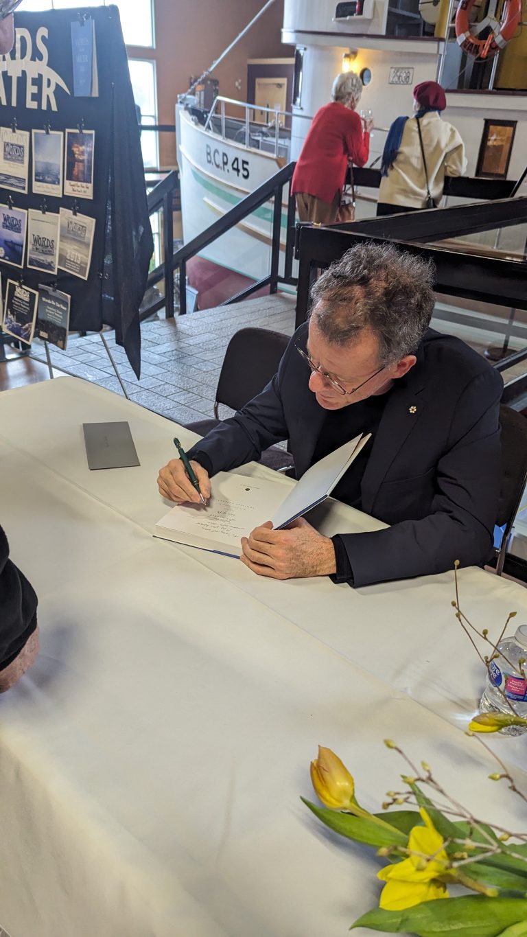 A man in a suit sits at a table signing a book, with a pen in hand. There are flowers on the table and people in the background, near a boat exhibit and a display board.