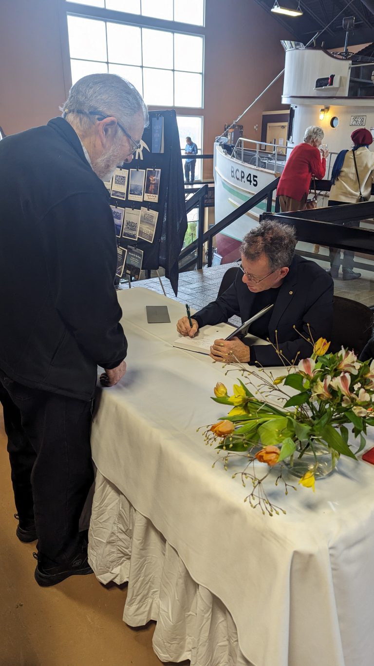A man sits at a table signing a book for another man standing in front of him. A large bouquet of flowers and display board are on the table, and a boat is visible in the background.