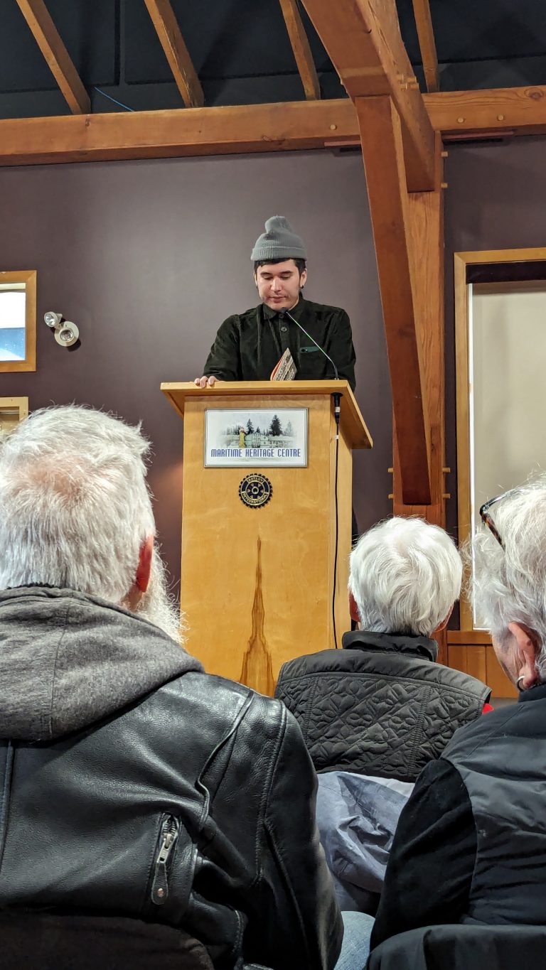 A person in a beanie speaks at a podium in front of an audience at the Mountain Nature Centre. The audience listens attentively in a wood-beamed room.